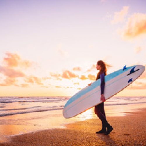 Surf girl holding surfboard at warm sunset or sunrise and ocean.